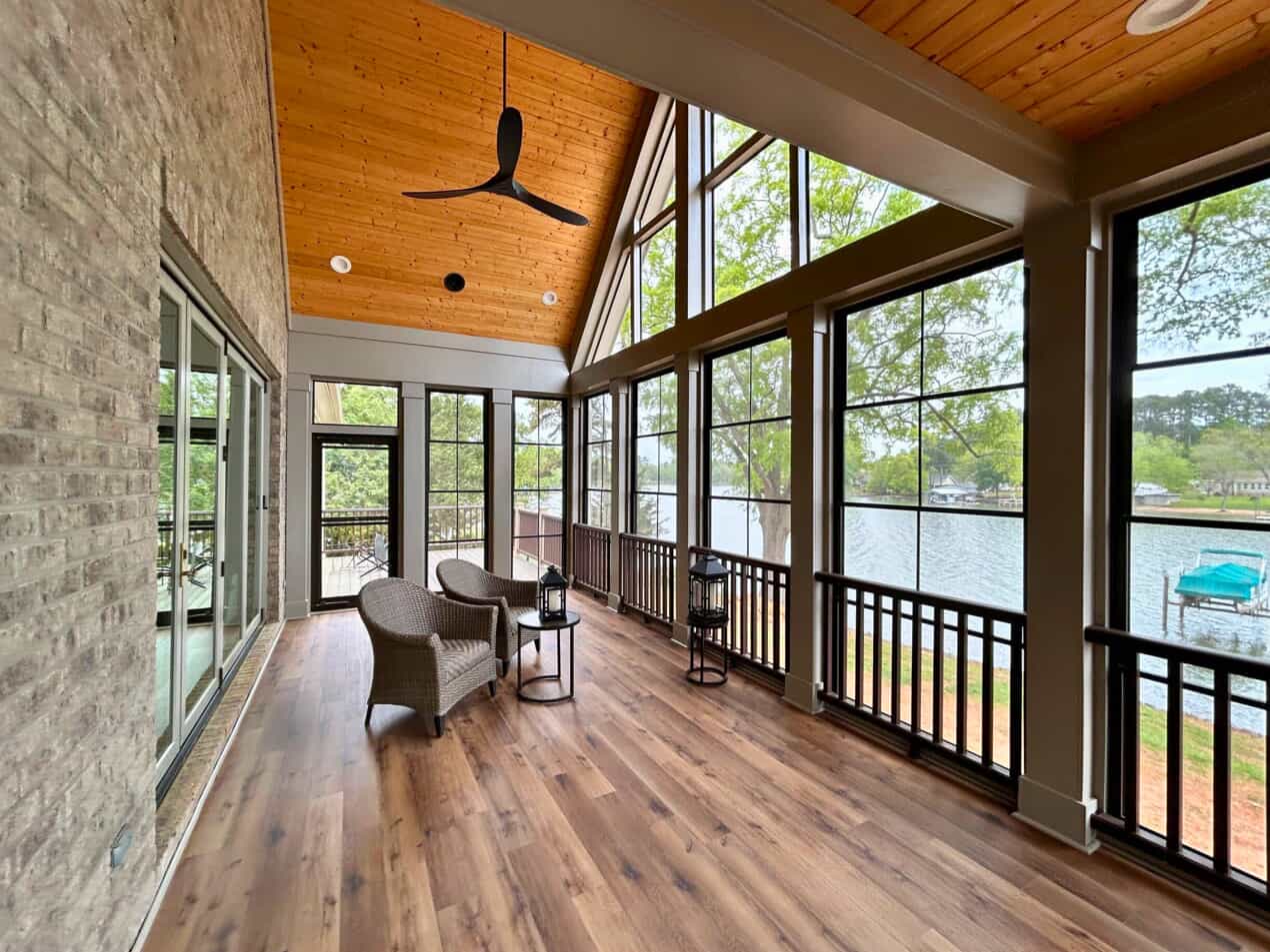 An expansive, bright screened-in porch with a high vaulted wood plank ceiling, a large black ceiling fan, and numerous windows offering a panoramic view of a lake and dock. Wicker chairs are arranged on a light wood-look floor.